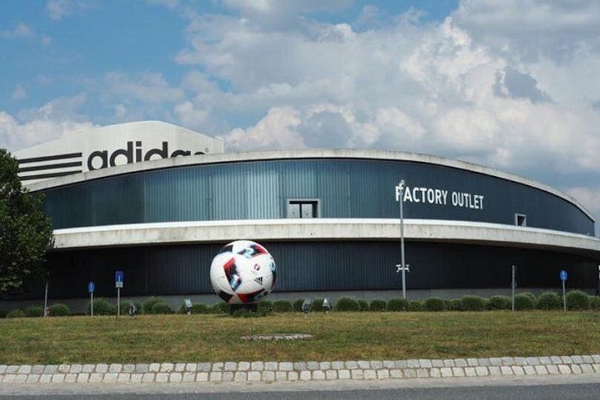 A modern building with a curved facade, a large Adidas logo, and a sign reading 'FACTORY OUTLET.' 1