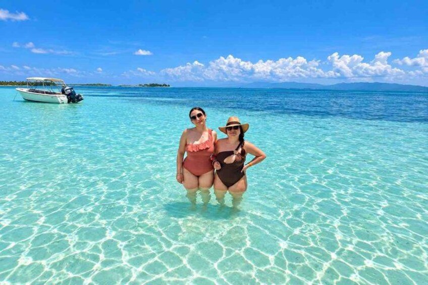 Deux personnes en maillot de bain, debout dans une eau turquoise et cristalline, avec un bateau en arrière-plan. 5