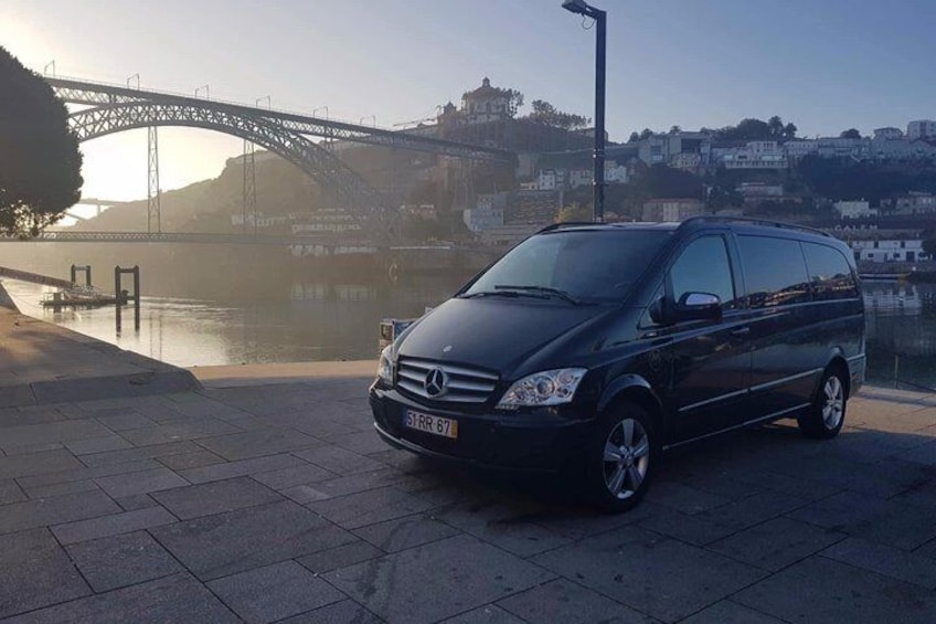 A black Mercedes-Benz van parked on a paved area near a river with a bridge in the background. 3