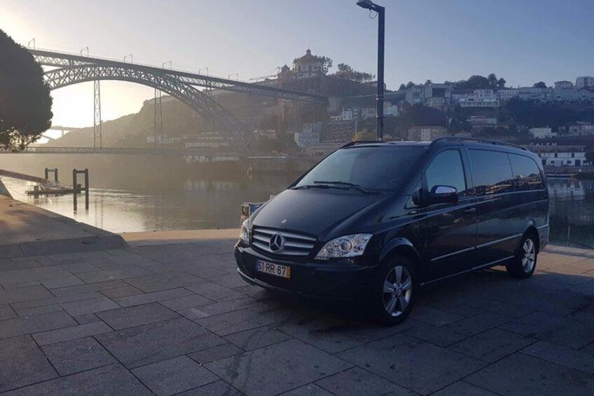 A black van parked on a paved area near a river with a bridge in the background. 3
