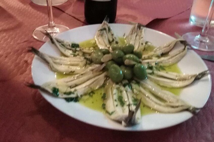 Une assiette de poisson grillé, garnie d’olives et d’herbes, accompagnée d’un verre de vin, posée sur une table. 3