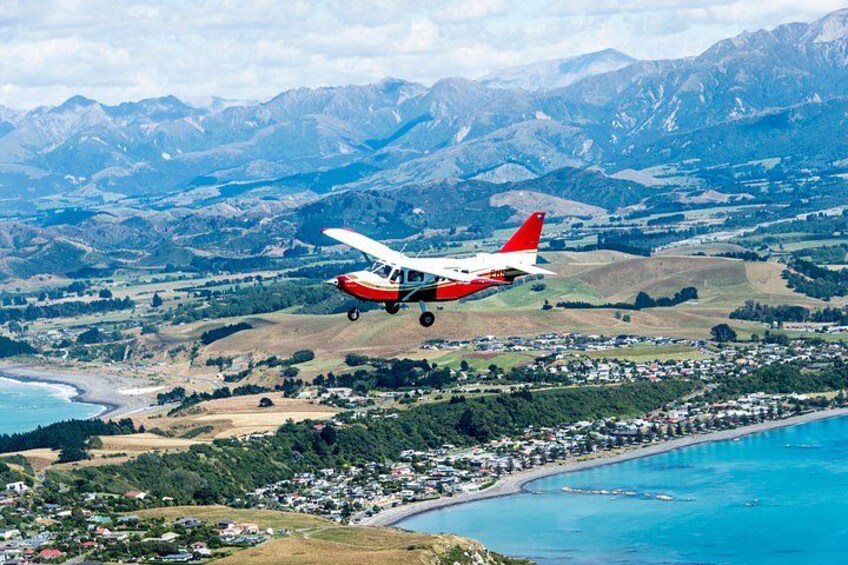 Una aeronave pequeña, roja y blanca volando sobre un pueblo costero con montañas al fondo. 3