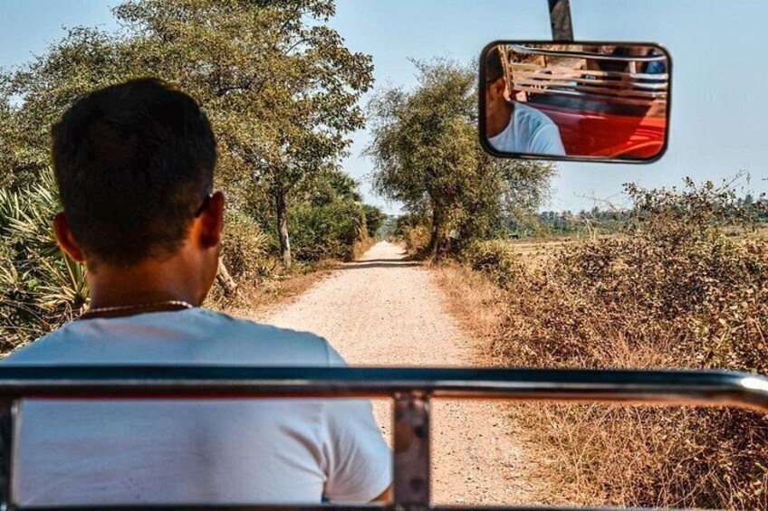 A person seated in a vehicle, looking out a side mirror at a rural road with trees and bushes. 4