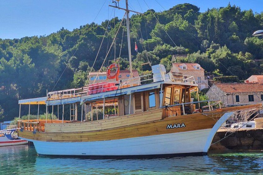 Un bote de madera llamado 'MARA' amarrado en un puerto con edificios y árboles al fondo. 4