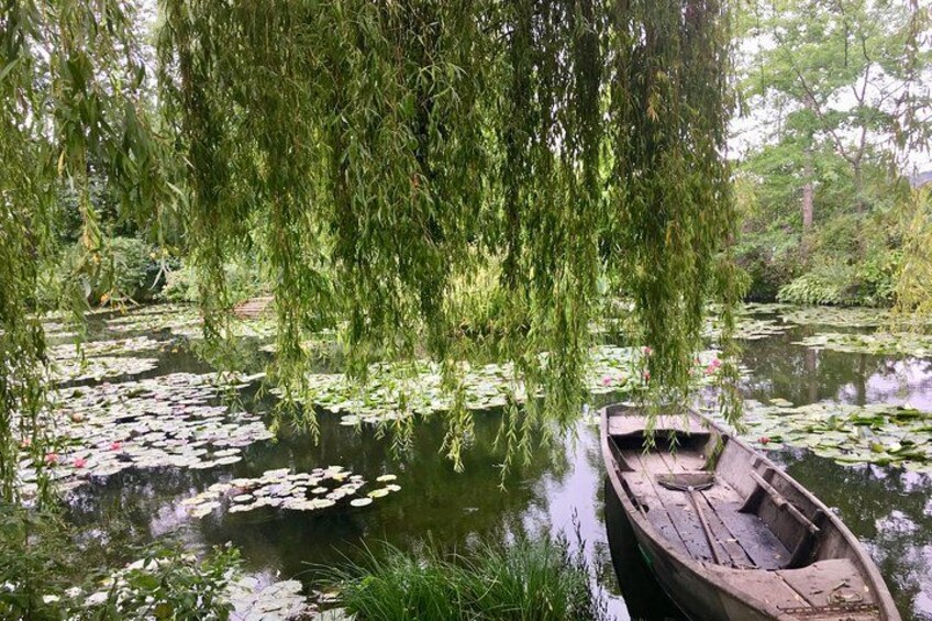 A wooden boat docked at a calm pond with lily pads and weeping willow branches overhanging. 3