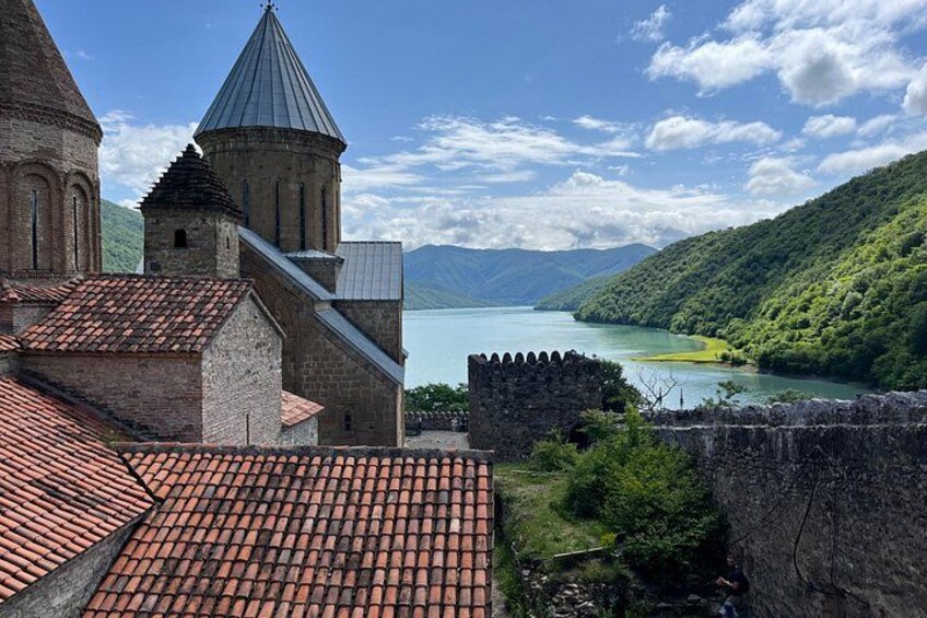Un edificio histórico con tejado de tejas rojas, una iglesia con campanario y un río con paisaje verde al fondo. 3