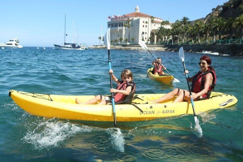 Deux personnes pratiquent le kayak sur une mer calme, avec un bâtiment situé sur la côte en arrière-plan. 1
