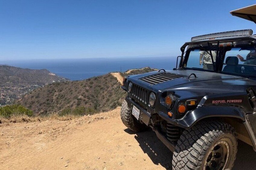 A Hummer with 'BIOFUEL POWERED' on the side parked on a dirt path with a view of the ocean and hills in the background. 2