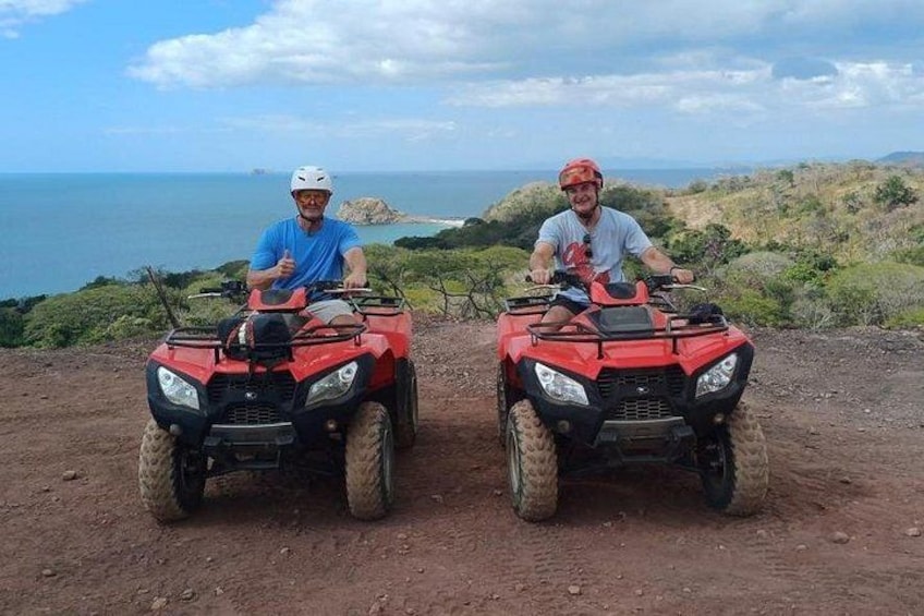 Two people on red ATVs on a dirt path with a view of the ocean and hills in the background. 1