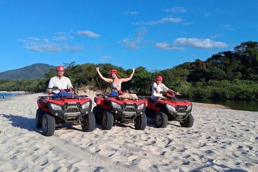 Three people on red ATVs on a sandy beach with a river and greenery in the background. 2
