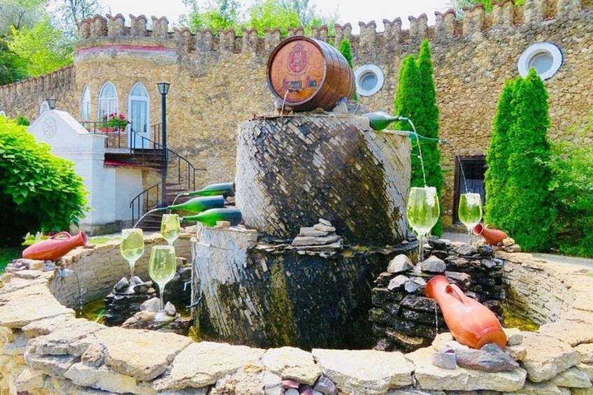 Entorno rústico al aire libre con una fuente de madera en forma de barril, rodeado de muros de piedra y follaje. 1