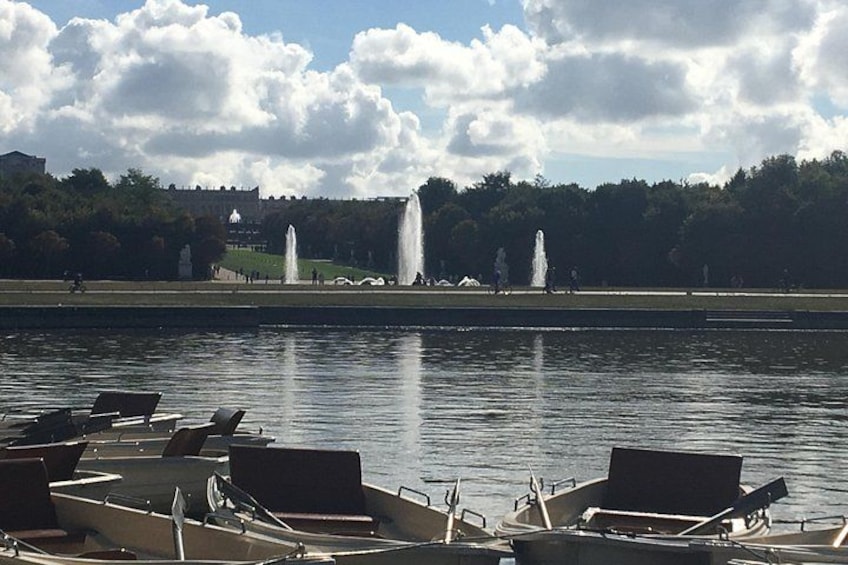 A calm lake with boats docked in the foreground and fountains in the background. 2