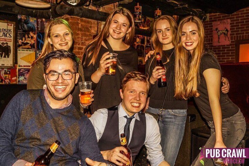 A group of people in a bar with drinks, a brick wall, and a poster. 1