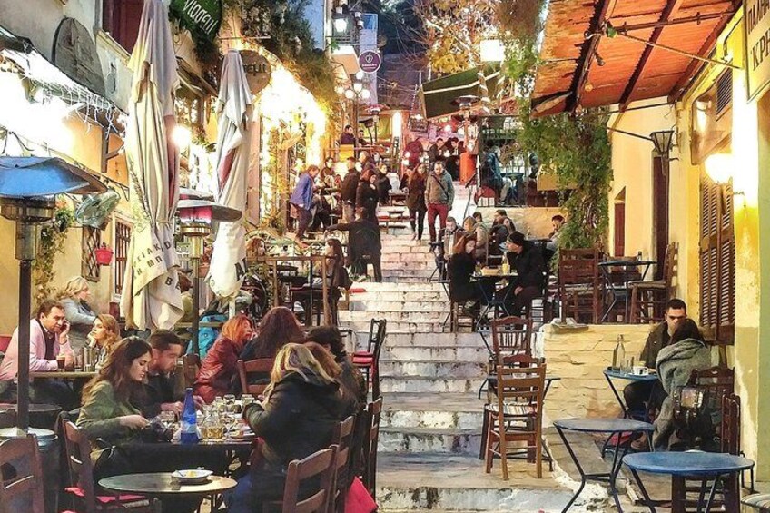 A bustling outdoor café with tables and chairs, a staircase leading up, and people walking. 1
