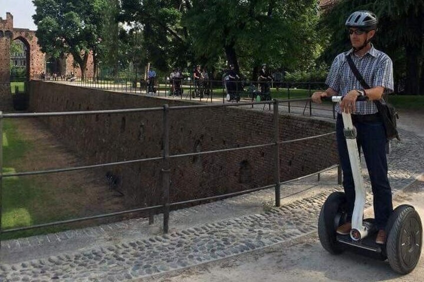 Eine Person fährt auf einem Segway auf einem befestigten Weg, im Hintergrund eine Ziegelmauer und viel Grün. 3