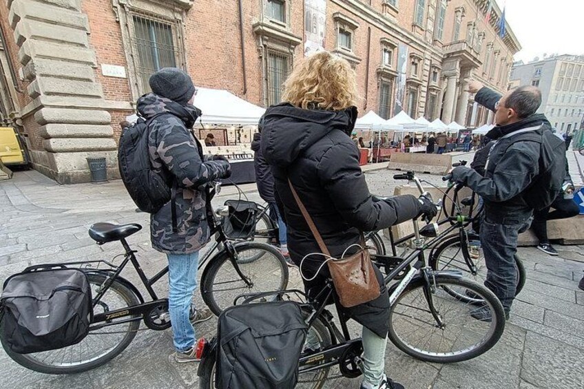 Folk står med cykler foran en murstensbygning, og udendørs siddepladser ses i baggrunden. 2