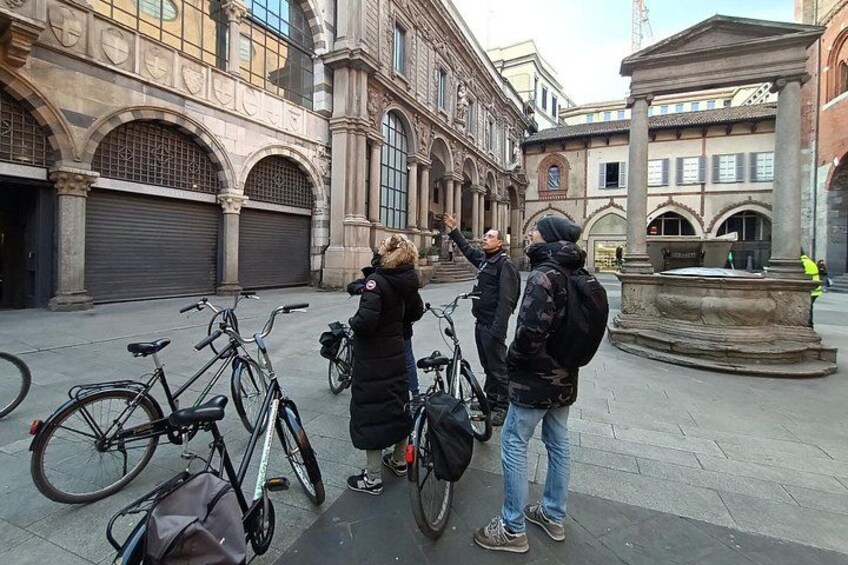 En gruppe mennesker står i en gårdsplads med cykler, historiske bygninger og en springvand. 3