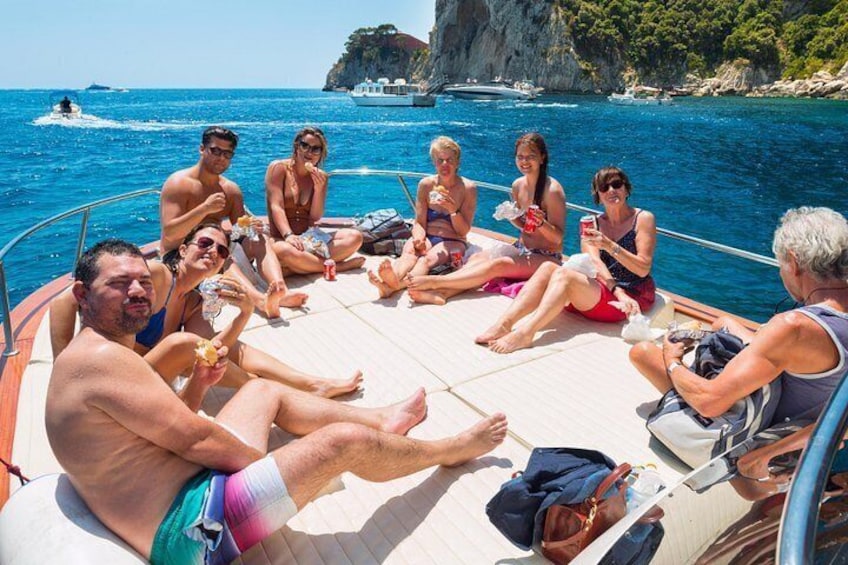 Un groupe de personnes se trouve sur un bateau, certaines tenant des boissons à la main, avec une côte rocheuse et une mer bleue et limpide en arrière-plan. 3
