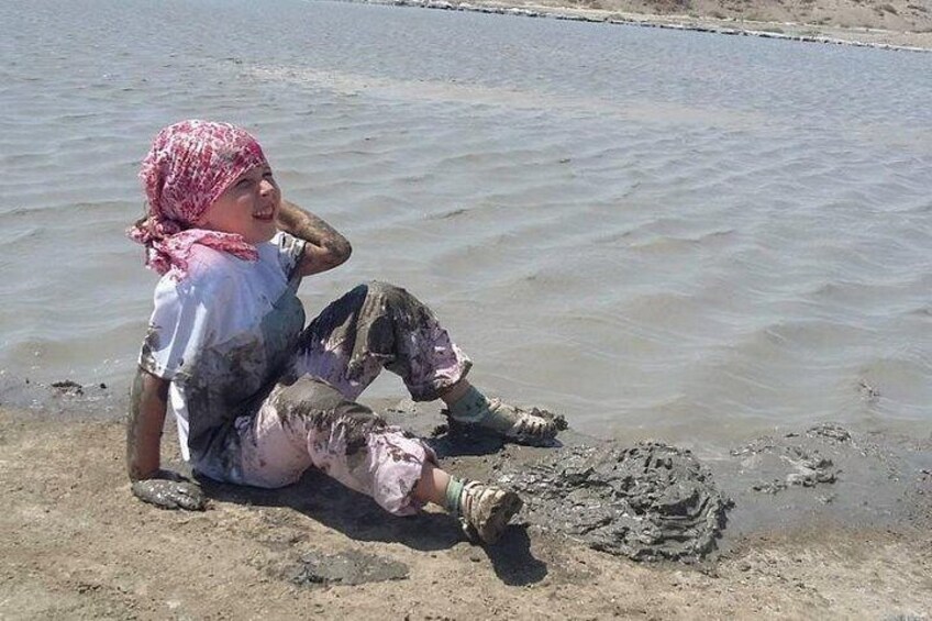A person sitting by a muddy water body, wearing a pink headscarf and muddy clothes. 4
