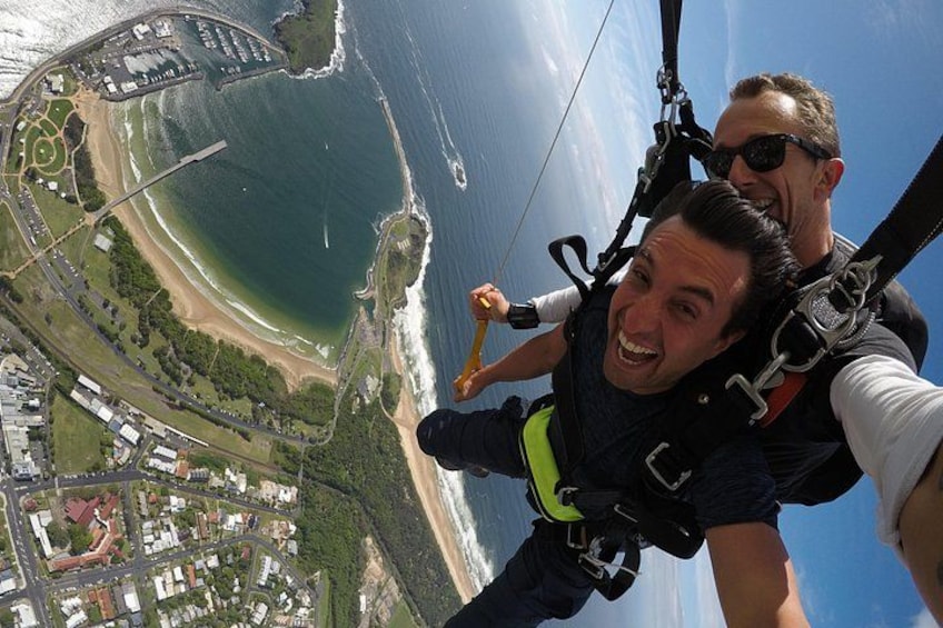 A person in a harness and helmet, with a parachute visible, and a coastal landscape with buildings and roads below. 3