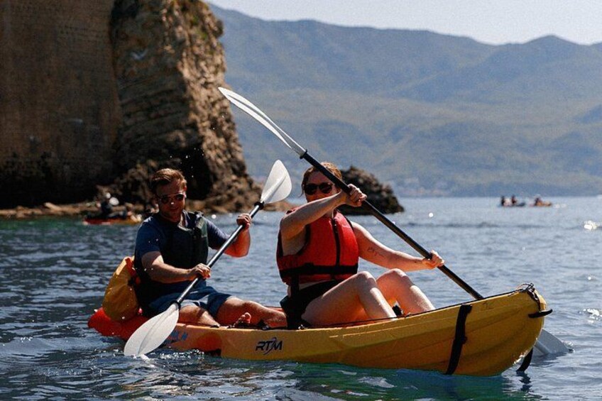 Two people kayaking on a calm body of water with a rocky coastline and mountains in the background. 1