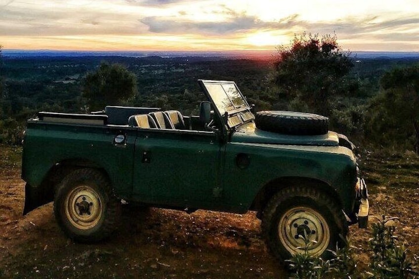 A green off-road vehicle parked on a dirt path with a sunset in the background. 1