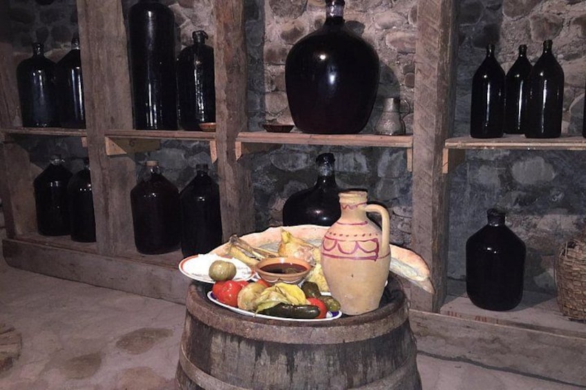 A rustic cellar with wooden shelves holding large black bottles, a wooden barrel with food items, and a decorative jug. 2