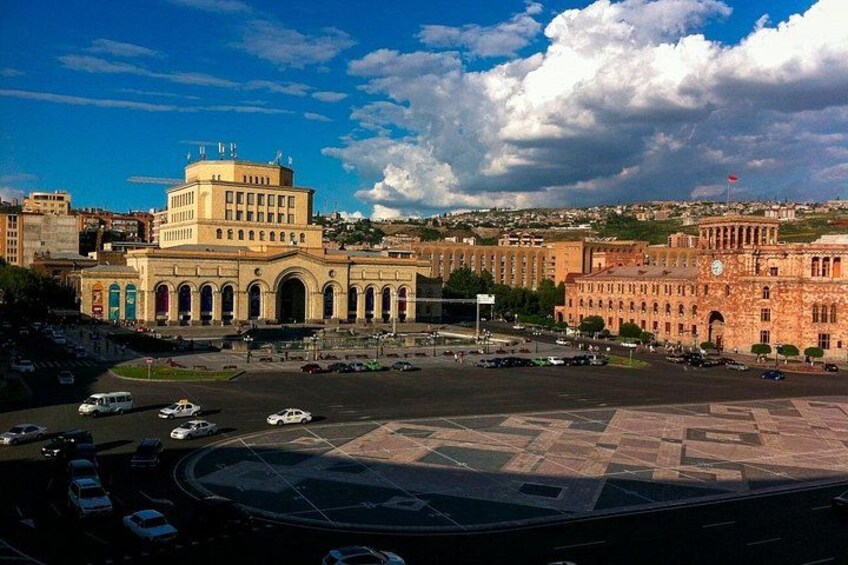 A cityscape with a prominent building with a dome and arched windows, a roundabout with vehicles, and a hilly background. 5