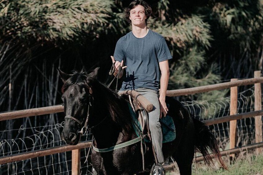 Una persona montando a caballo en un entorno al aire libre, con una cerca de madera y árboles al fondo. 4