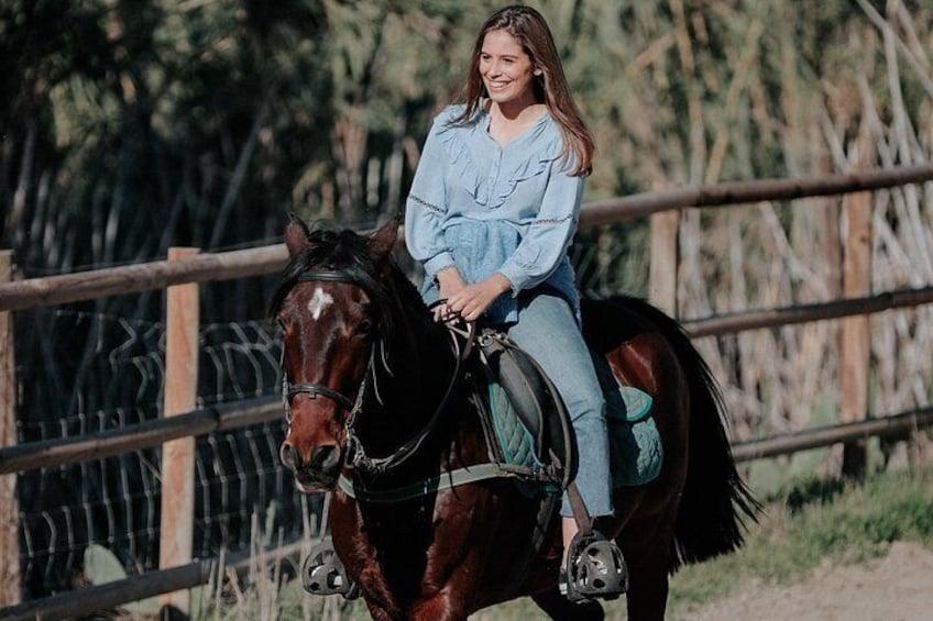 Una persona montando a caballo en un entorno al aire libre, con una cerca de madera y árboles al fondo. 3