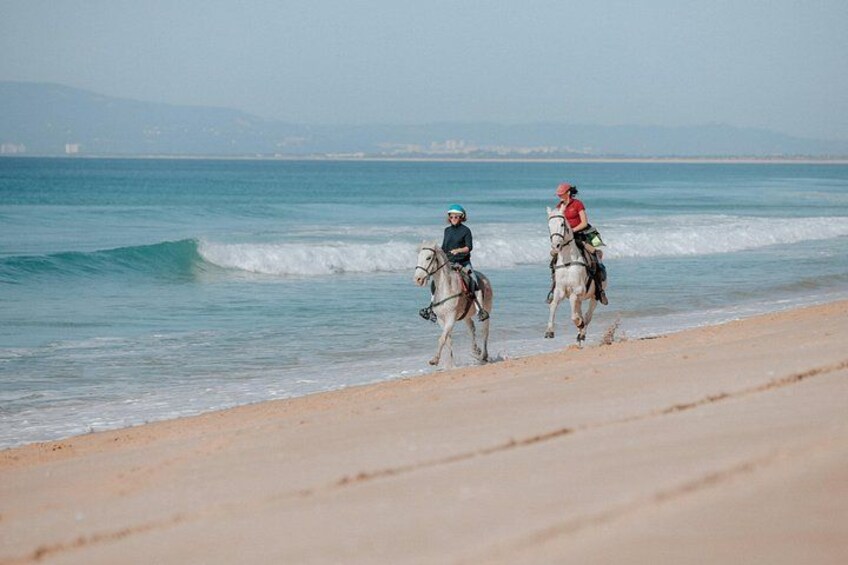 Two people riding horses along a beach with waves in the background. 2