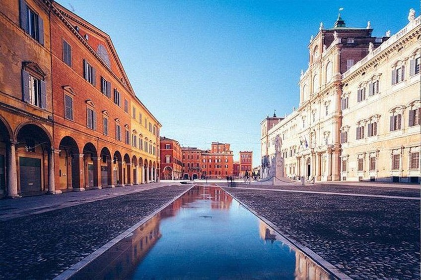 A cobblestone street with historic buildings on either side, a clear sky, and a reflective water surface. 4