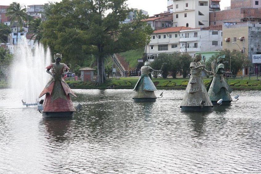 Una estatua de una mujer con vestido junto a una fuente en un parque, con edificios al fondo. 3