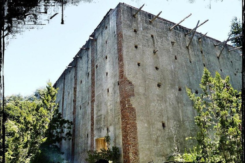 A large, weathered concrete building with a brick section and multiple small windows, surrounded by greenery. 4