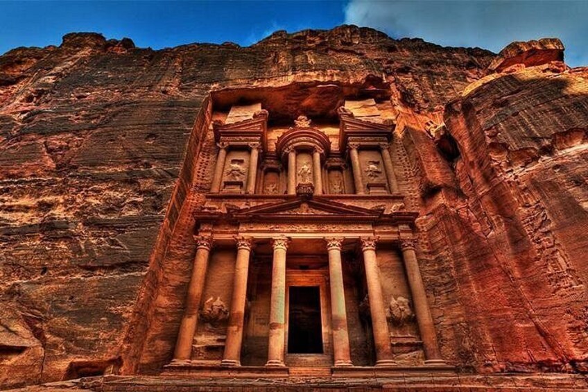 Una gran estructura excavada en la roca, con columnas y un acceso central, frente a un paisaje montañoso. 2