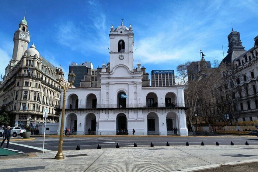 Un edificio histórico con una torre de reloj, rodeado de edificios modernos y un cielo despejado. 2