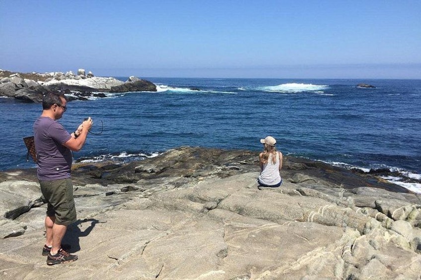 A rocky coastline with waves crashing onto the shore, a person taking a photo, and another person sitting and looking at the ocean. 4