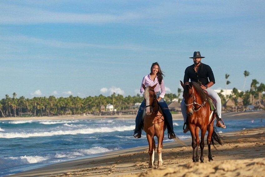 Two people riding horses on a beach with ocean waves and palm trees in the background. 2