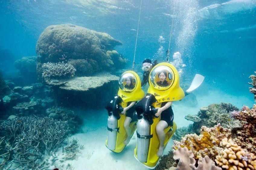 Dos personas con equipo de buceo amarillo bajo el agua, rodeadas de arrecifes de coral y vida marina. 1