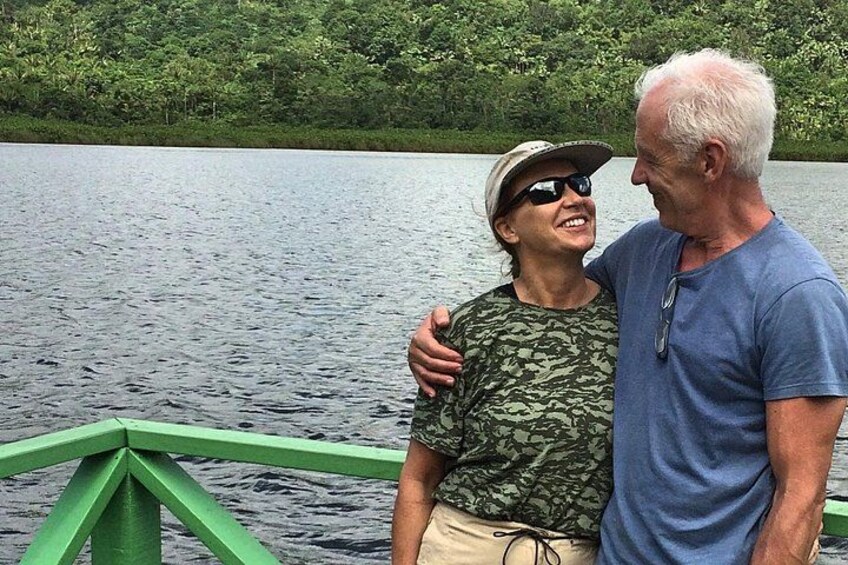 Two people on a boat with a green railing, surrounded by a calm lake and dense greenery. 2