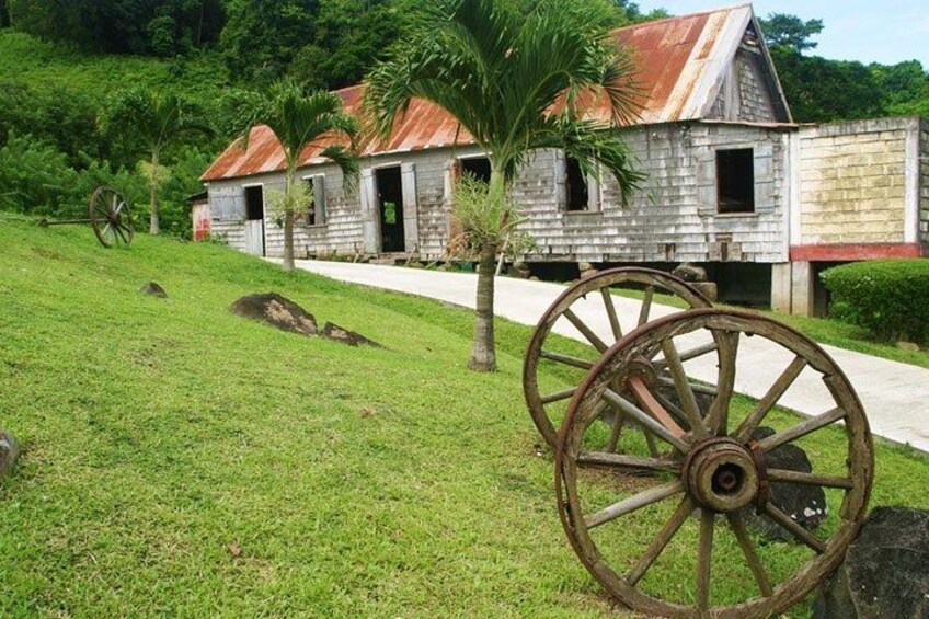 A rustic wooden building with a rusted roof, a palm tree, and a large wooden wheel in a grassy area. 2