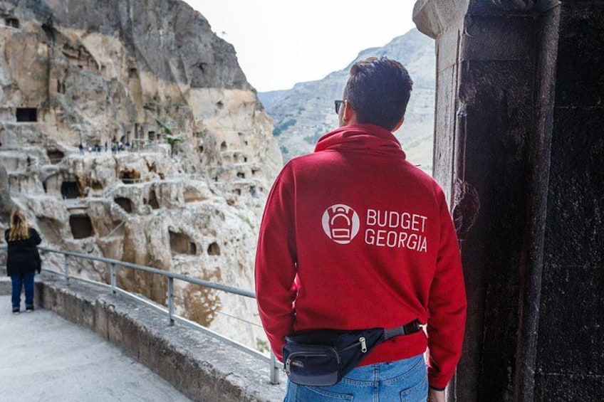 A person in a red jacket with 'BUDGET GEORGIA' on the back, standing by a rocky cliff with ancient cave dwellings. 1