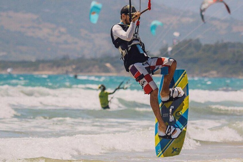Una persona practicando kiteboarding en una tabla con un diseño colorido, en una zona costera con olas y otros kiteboarders al fondo. 2