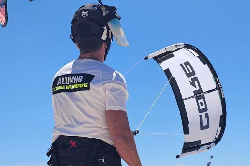 Una persona con un'imbracatura da paracadute e un casco, indossa una maglietta bianca con la scritta 'ALUMNO' e 'EXPORA WATERSPORTS' sulla schiena. 1