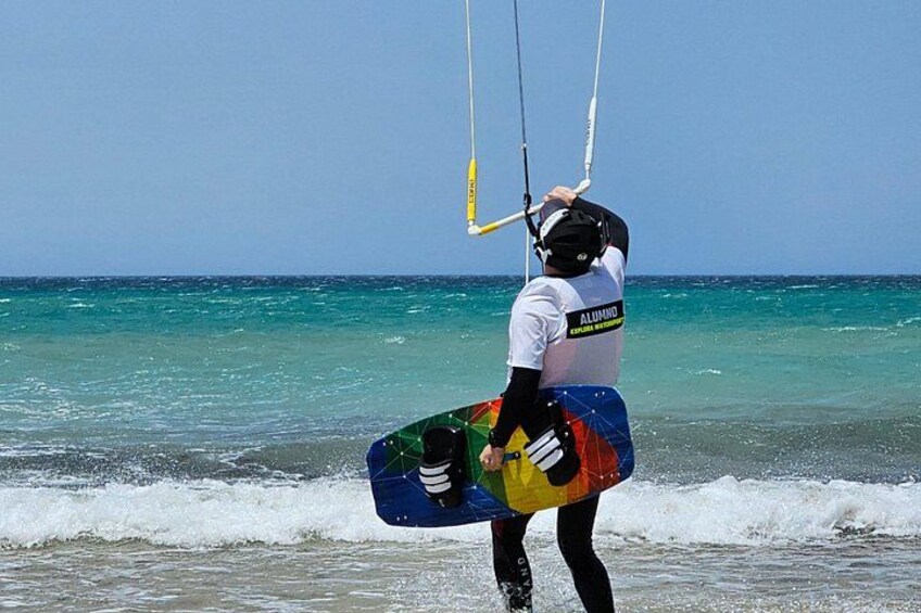 Una persona con una tavola a vela colorata cammina sulla spiaggia, tenendo in mano un aquilone e indossando un casco. 4