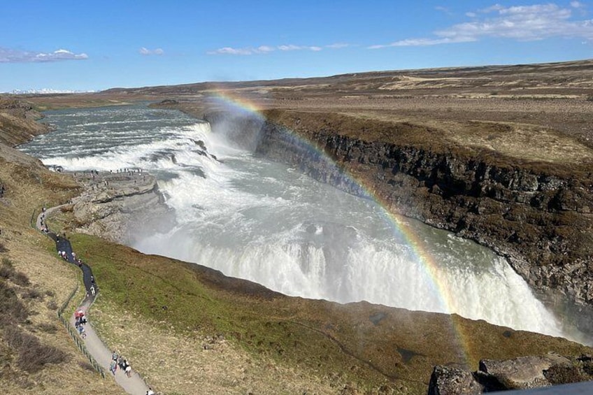 A waterfall with a rainbow, a paved path, and a distant mountain range. 2