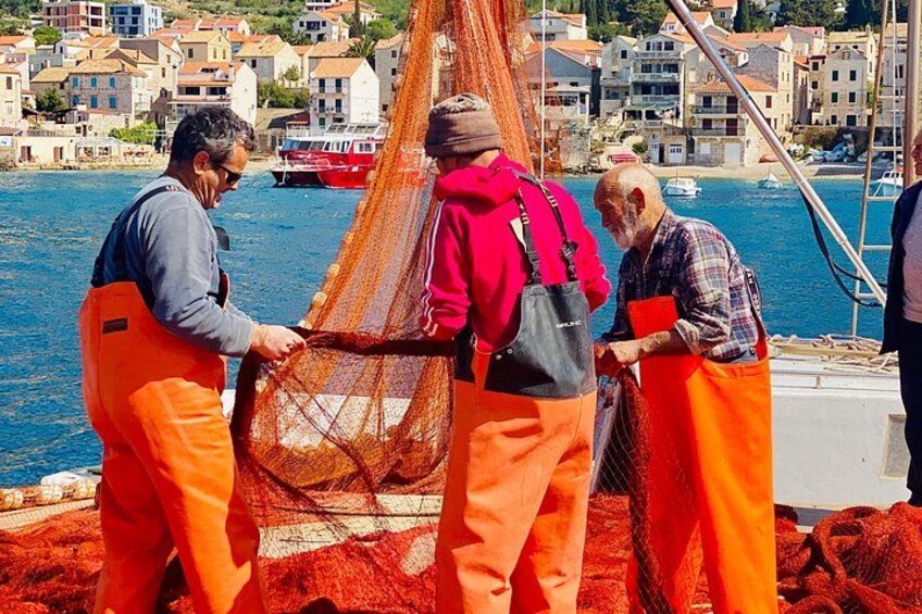 Personer hanterar ett stort orangefärgat fisknät på en brygga, med byggnader och båtar i bakgrunden. 4