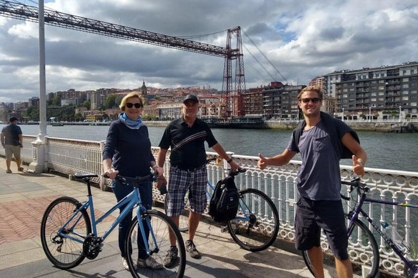 Three people standing by a waterfront with bicycles, a bridge, and buildings in the background. 1