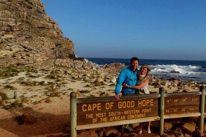 A couple standing by a sign at Cape of Good Hope, with the ocean and rocky cliffs in the background. 1
