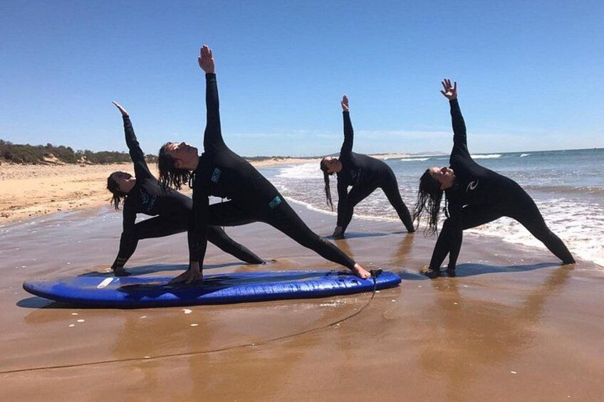 Fire personer i sorte våddragter laver yoga på en strand med en blå surfbræt. 1